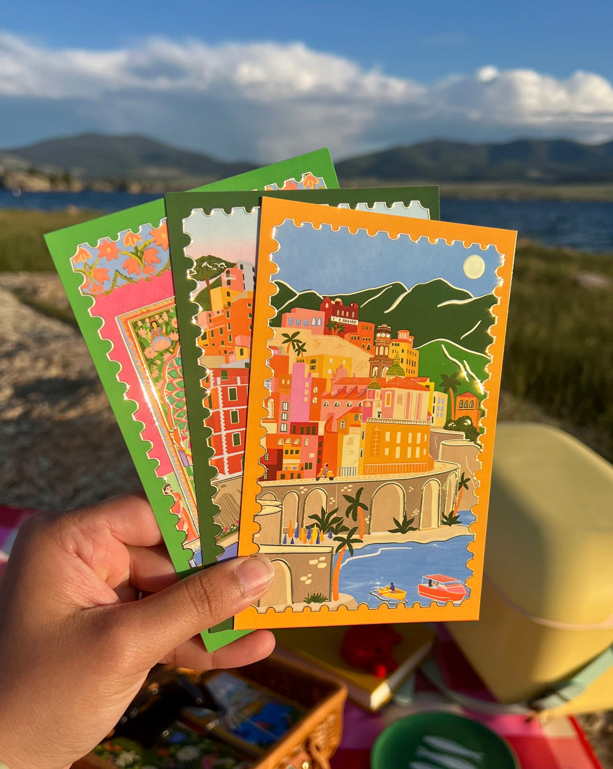 Hand holding a fan of postcards including Under the Amalfi Sun, lake and mountains in background