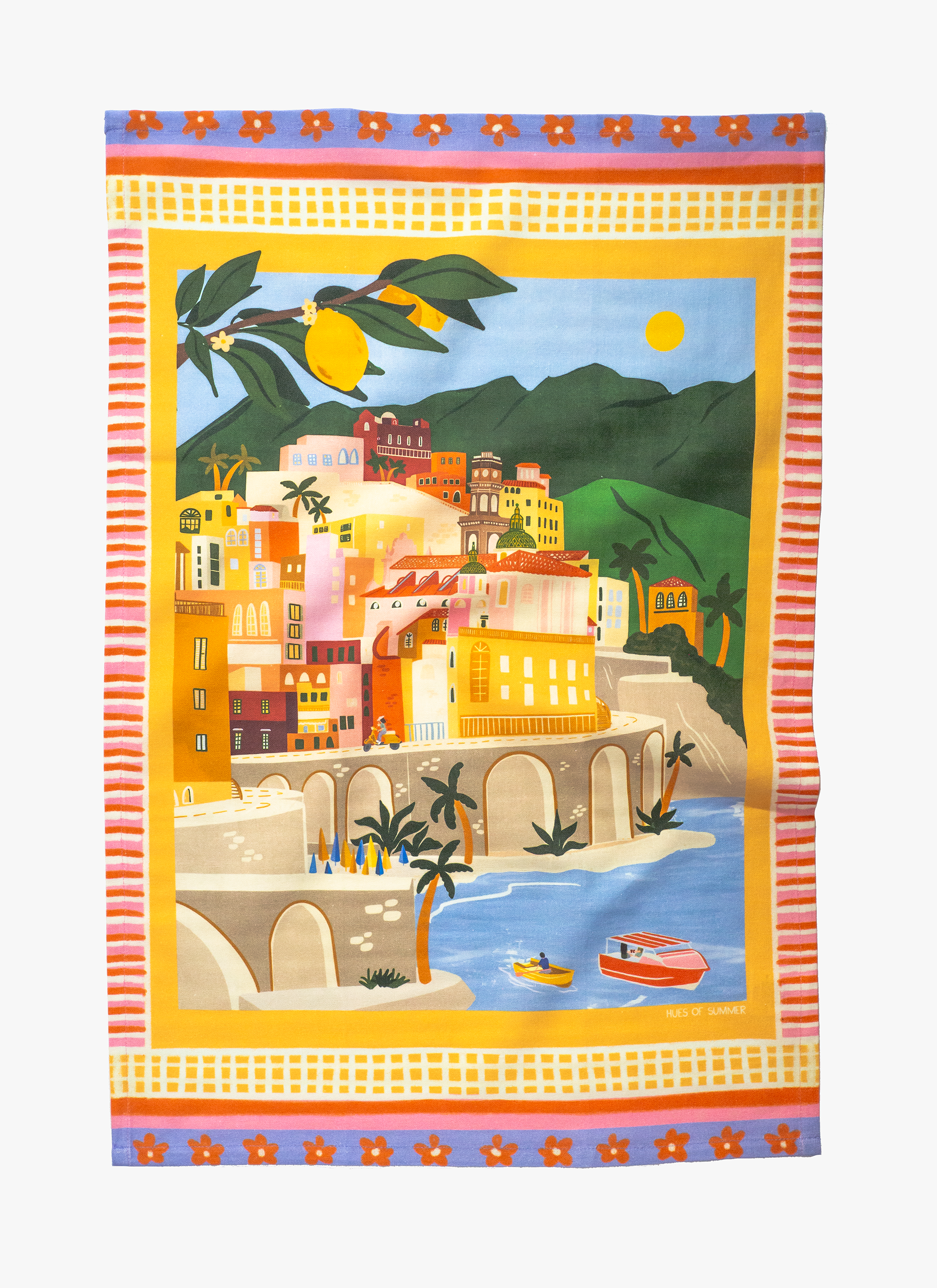A colorful tea towel featuring a vibrant illustration of a coastal town with lemon trees and a blue sky.