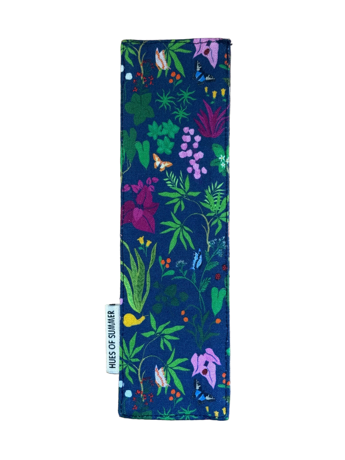 A rectangular fabric bookmark with a colorful botanical print on a navy blue background, laid flat on a light brown wooden surface. A small white fabric tag on the left side reads &