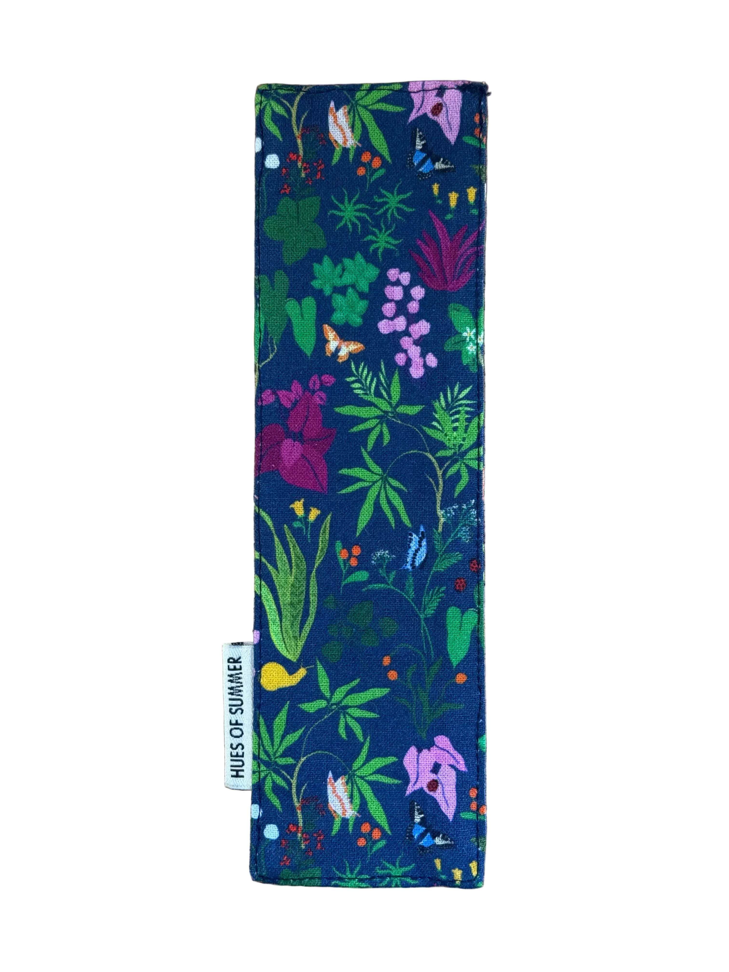 A rectangular fabric bookmark with a colorful botanical print on a navy blue background, laid flat on a light brown wooden surface. A small white fabric tag on the left side reads &