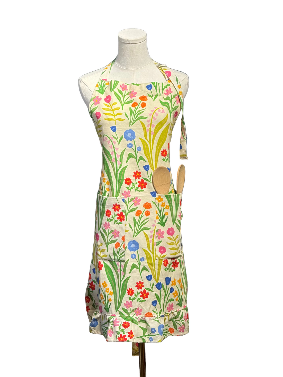 A colorful floral apron displayed on a white mannequin torso, featuring a cream base with a vibrant print of wildflowers and green leaves, with two wooden spoons tucked into the front pocket, set against a blue paneled wall.