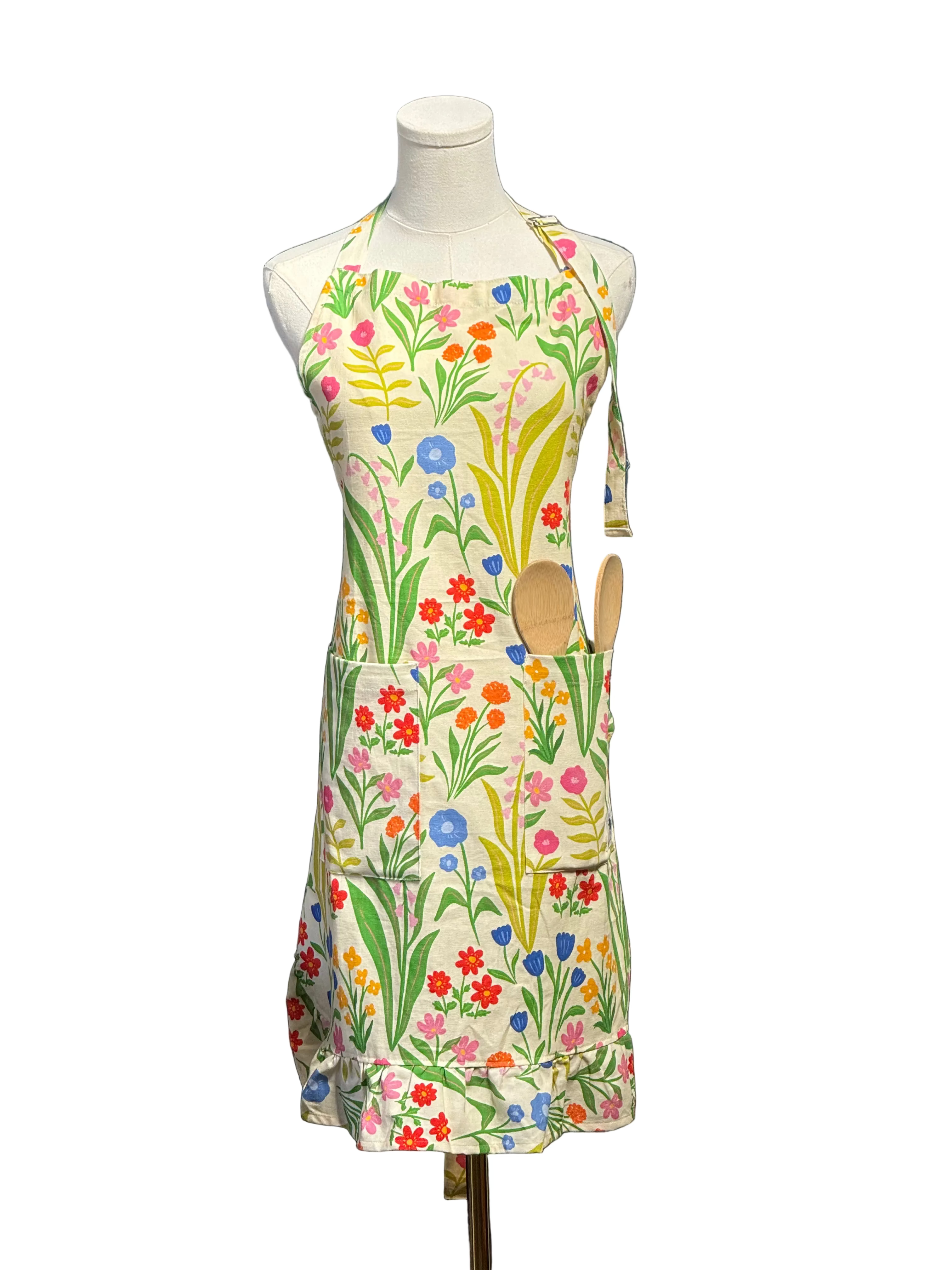 A colorful floral apron displayed on a white mannequin torso, featuring a cream base with a vibrant print of wildflowers and green leaves, with two wooden spoons tucked into the front pocket, set against a blue paneled wall.