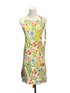 A colorful floral apron displayed on a white mannequin torso, featuring a cream base with a vibrant print of wildflowers and green leaves, with two wooden spoons tucked into the front pocket, set against a blue paneled wall.