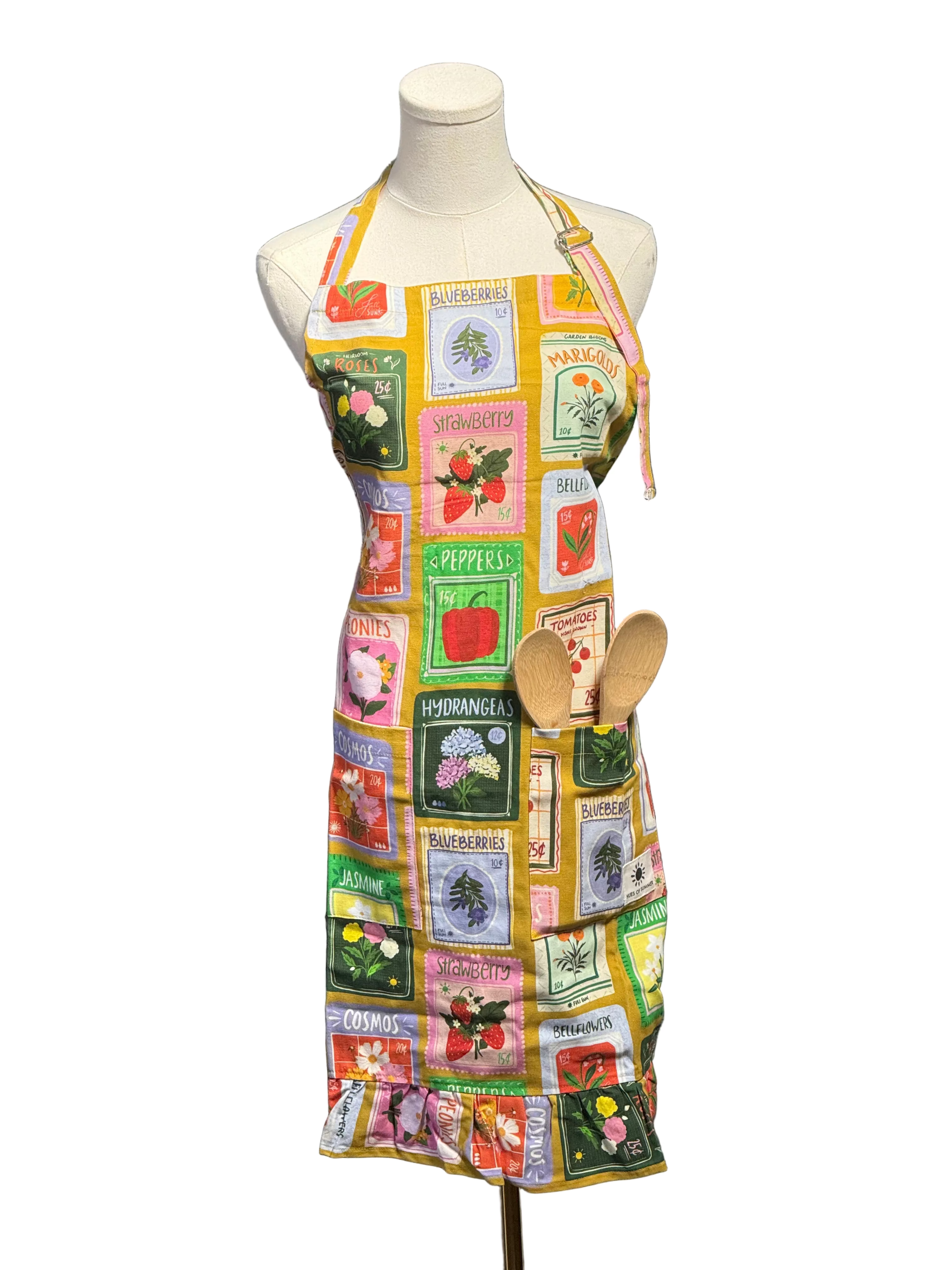A multicolor apron with a vintage fruit-and-vegetable patchwork print is displayed on a white mannequin dress form. The apron features a yellow border and includes a front pocket holding two wooden spoons, set against a blue paneled wall background.