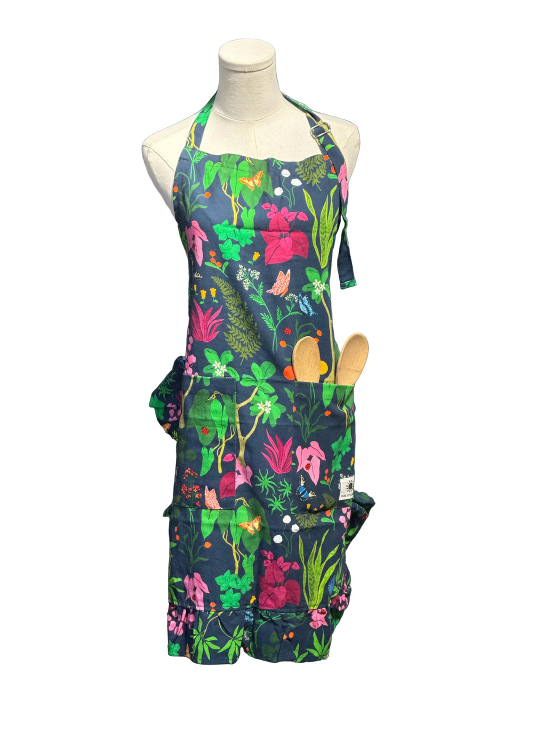 A colorful garden-themed apron with a dark navy background and a vibrant floral print, displayed on a mannequin torso. The apron features pockets containing gardening tools and is set against a blue paneled wall.