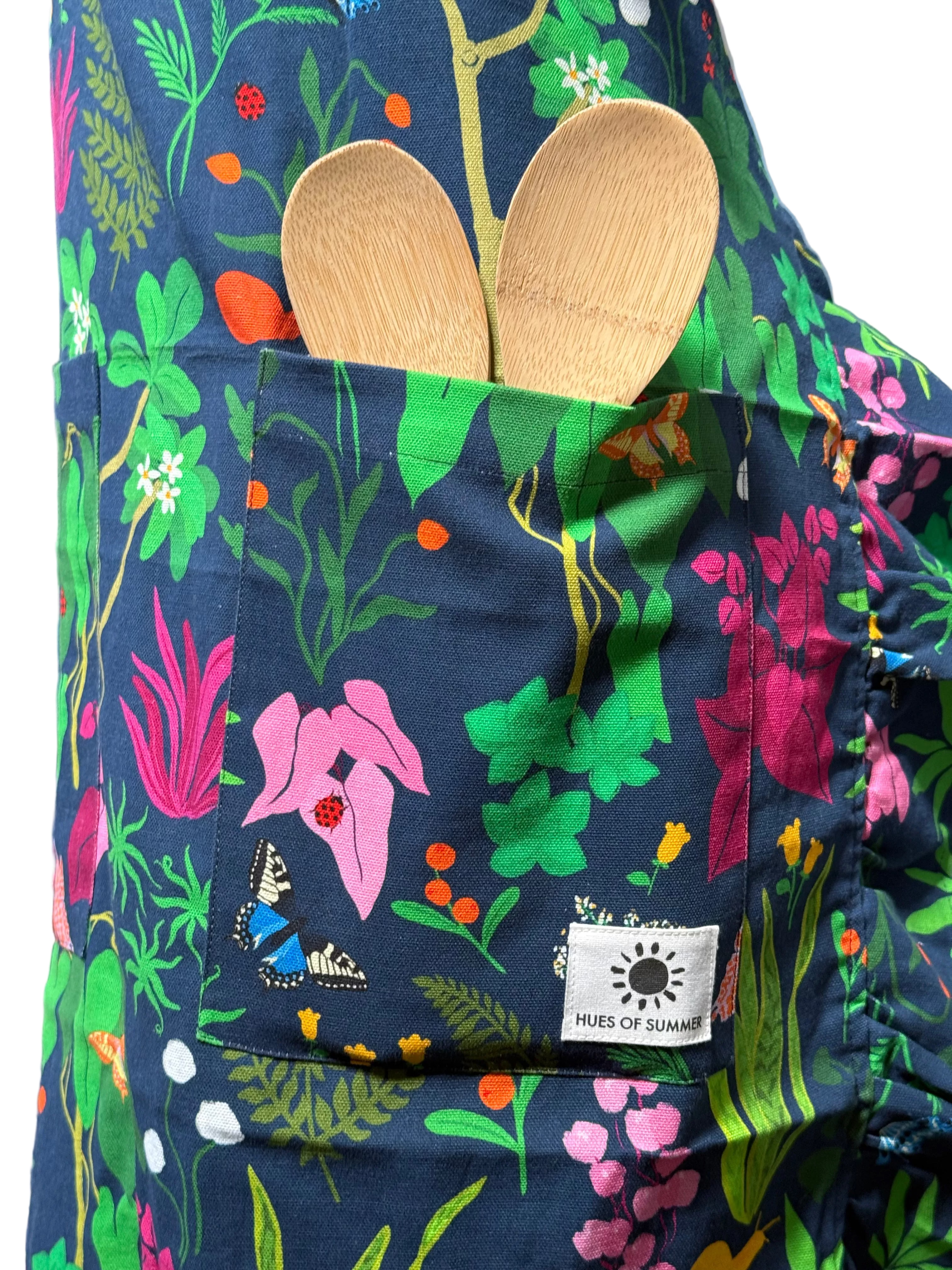 A close-up of a dark navy apron with a vibrant, multicolored botanical and floral print. A front pocket holds two light wood spoons, and a white sewn-on label reading &