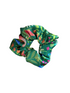 A green fabric scrunchie with a tropical print featuring pink flamingos, orange flowers, and green palm leaves, resting on a light brown wooden surface.