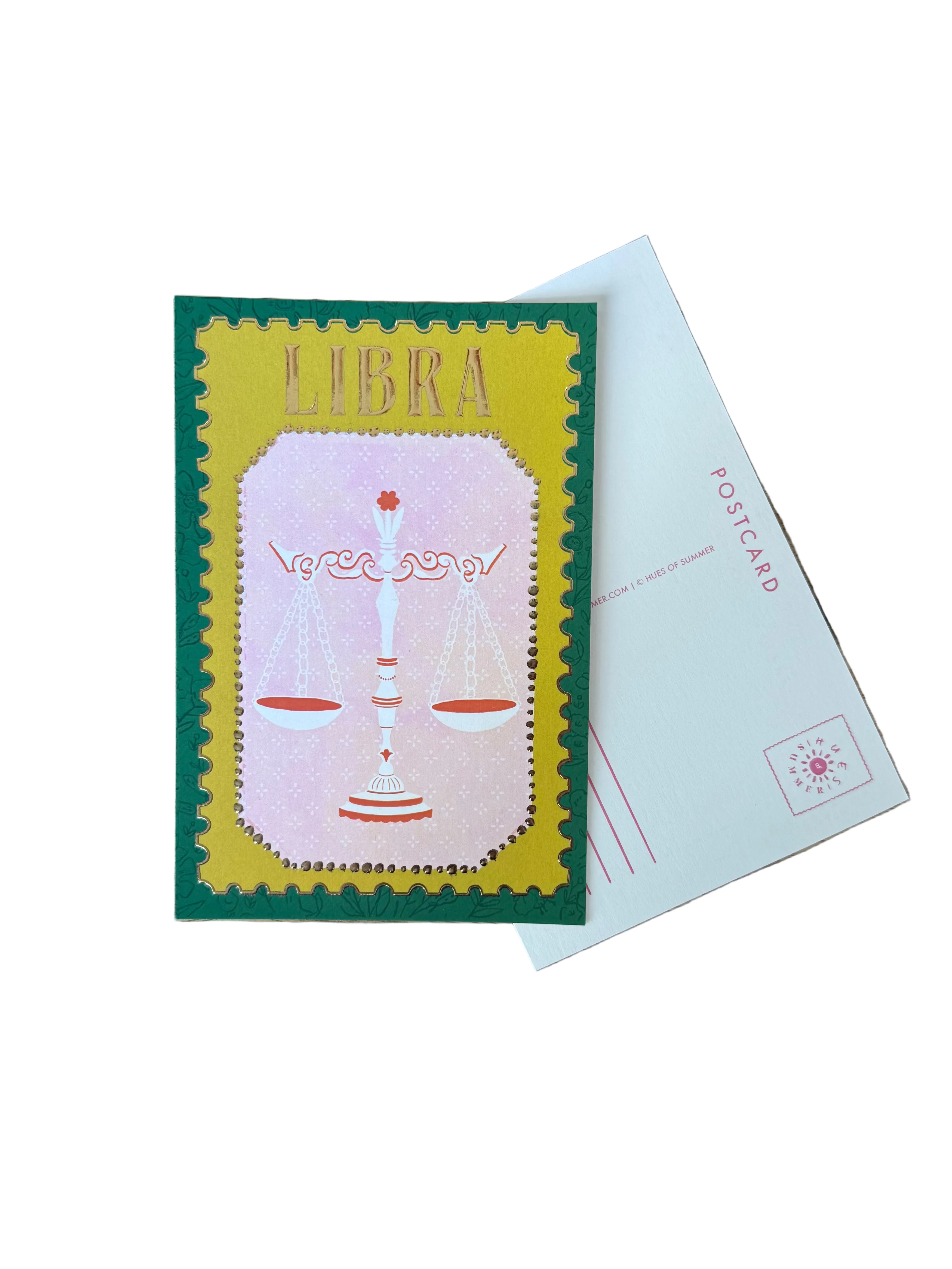 A rectangular art print or postcard featuring a pink and yellow illustration of a scale of justice, placed on a wooden table next to a white postcard.