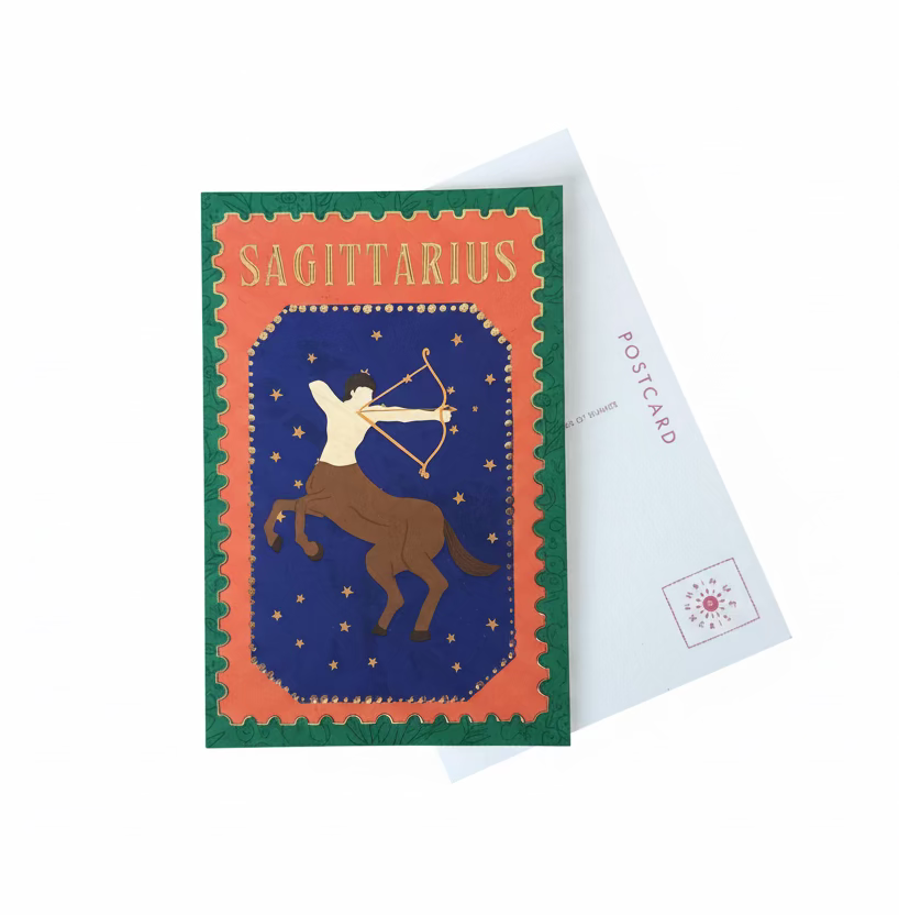 A Sagittarius zodiac-themed postcard featuring a graphic illustration of a rider on a horse under a starry sky, displayed on a wooden table next to a white postcard envelope.